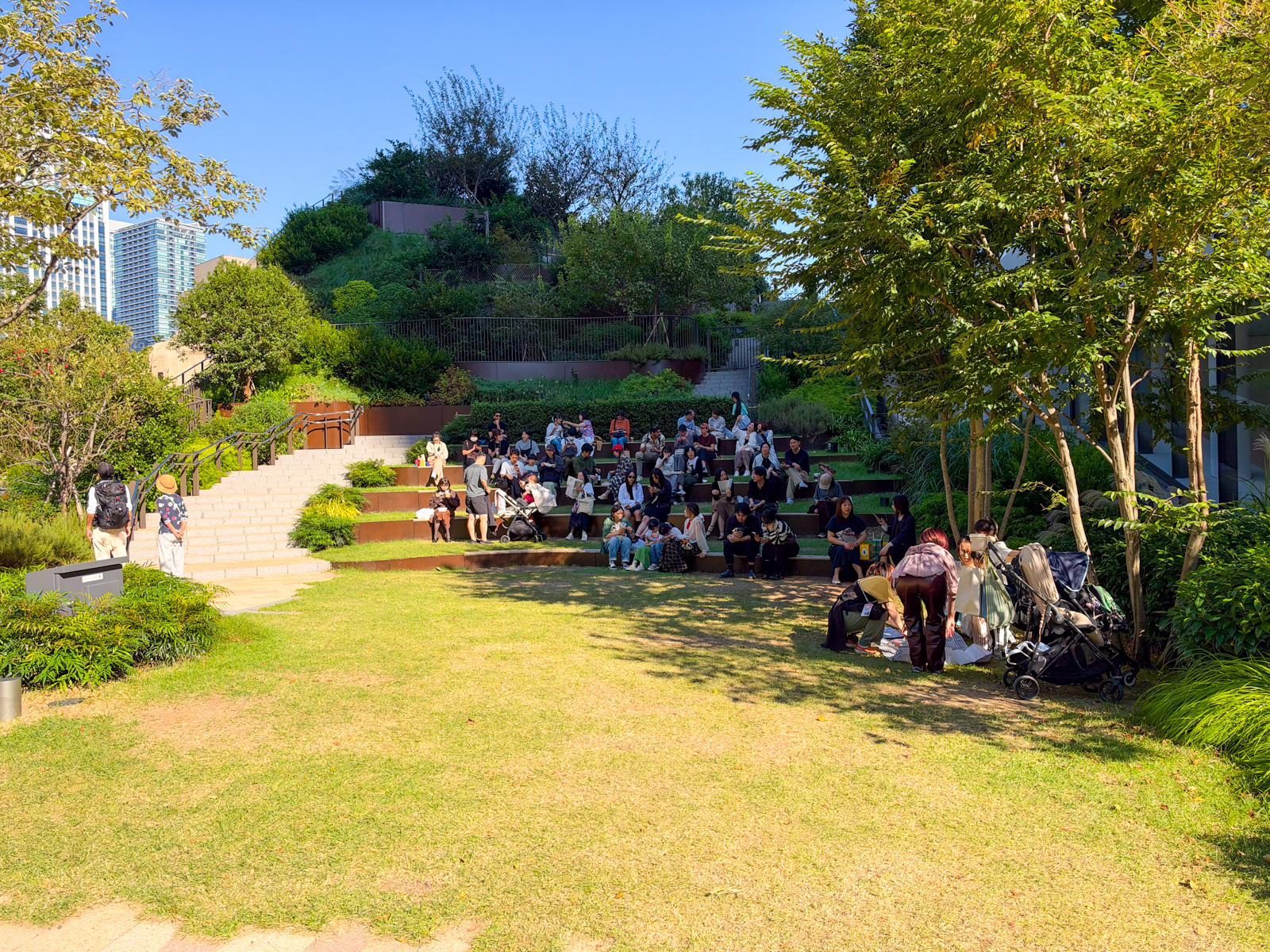 広場。段状の芝生に大勢の人が腰を下ろしている。子ども連れ、カップル、一人でいる人。緑の段丘と木立に囲まれた昼下がり