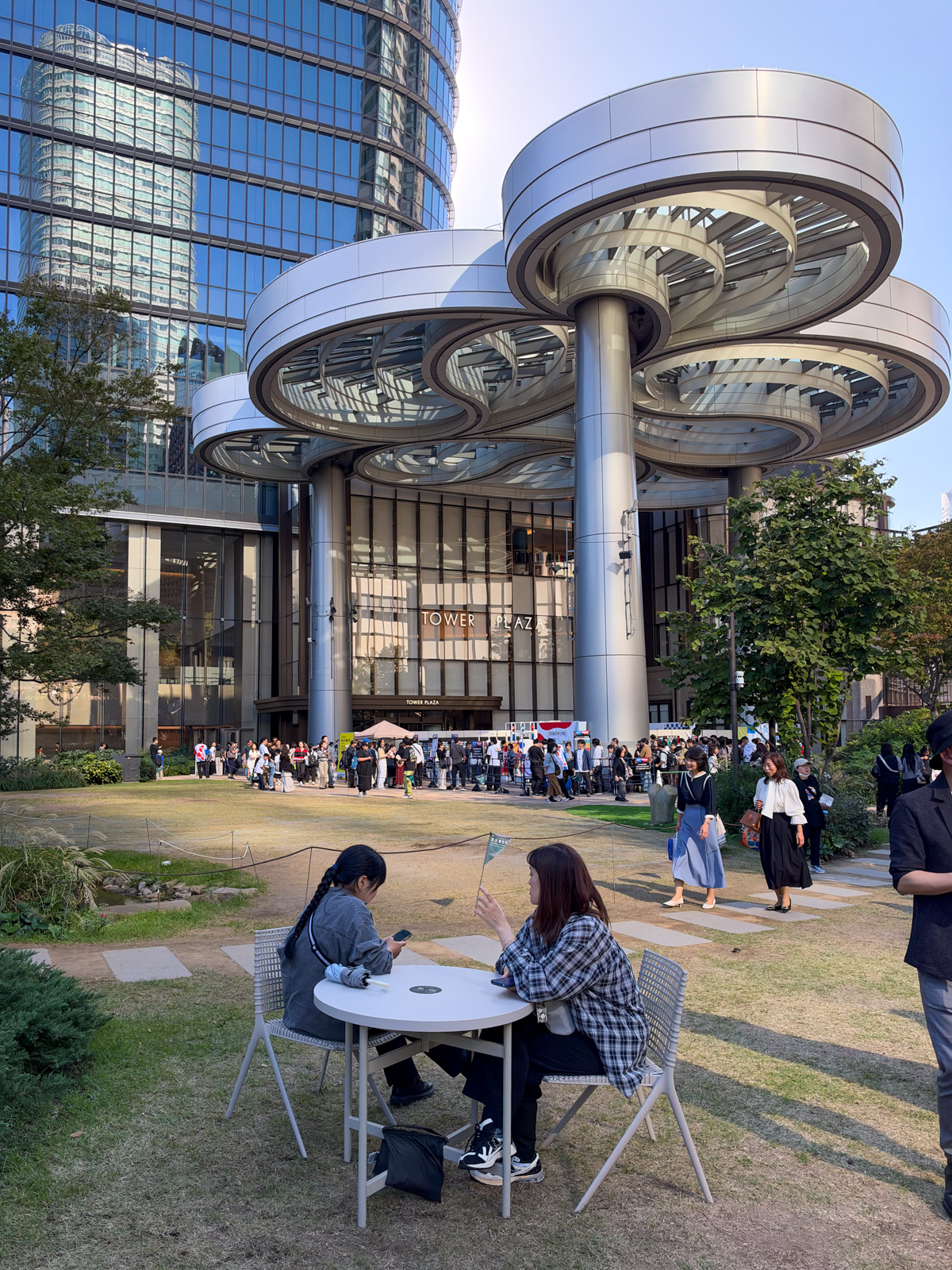 Tower Plazaの円盤型カノピー。直径約18mの水平な円盤が柱の上に3枚積み重なり、その下の芝生にマーケットが立ち、大勢の人が集まっている