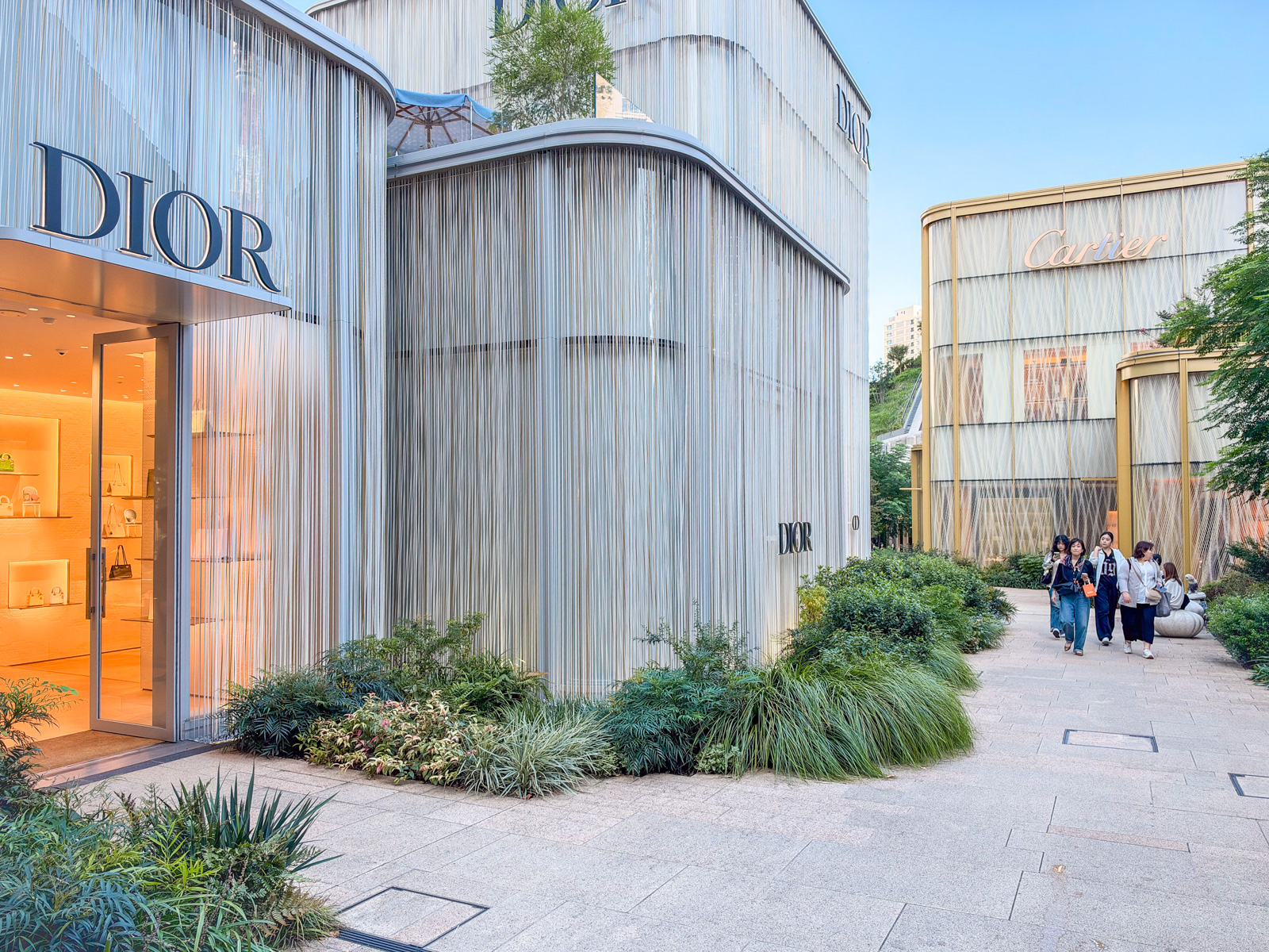 DiorとCartierのブランドパビリオンが並ぶ通路。左に銀色のステンレスロッドカーテン(Dior)、右奥に金色のフレーム(Cartier)。植栽が道を縁どる