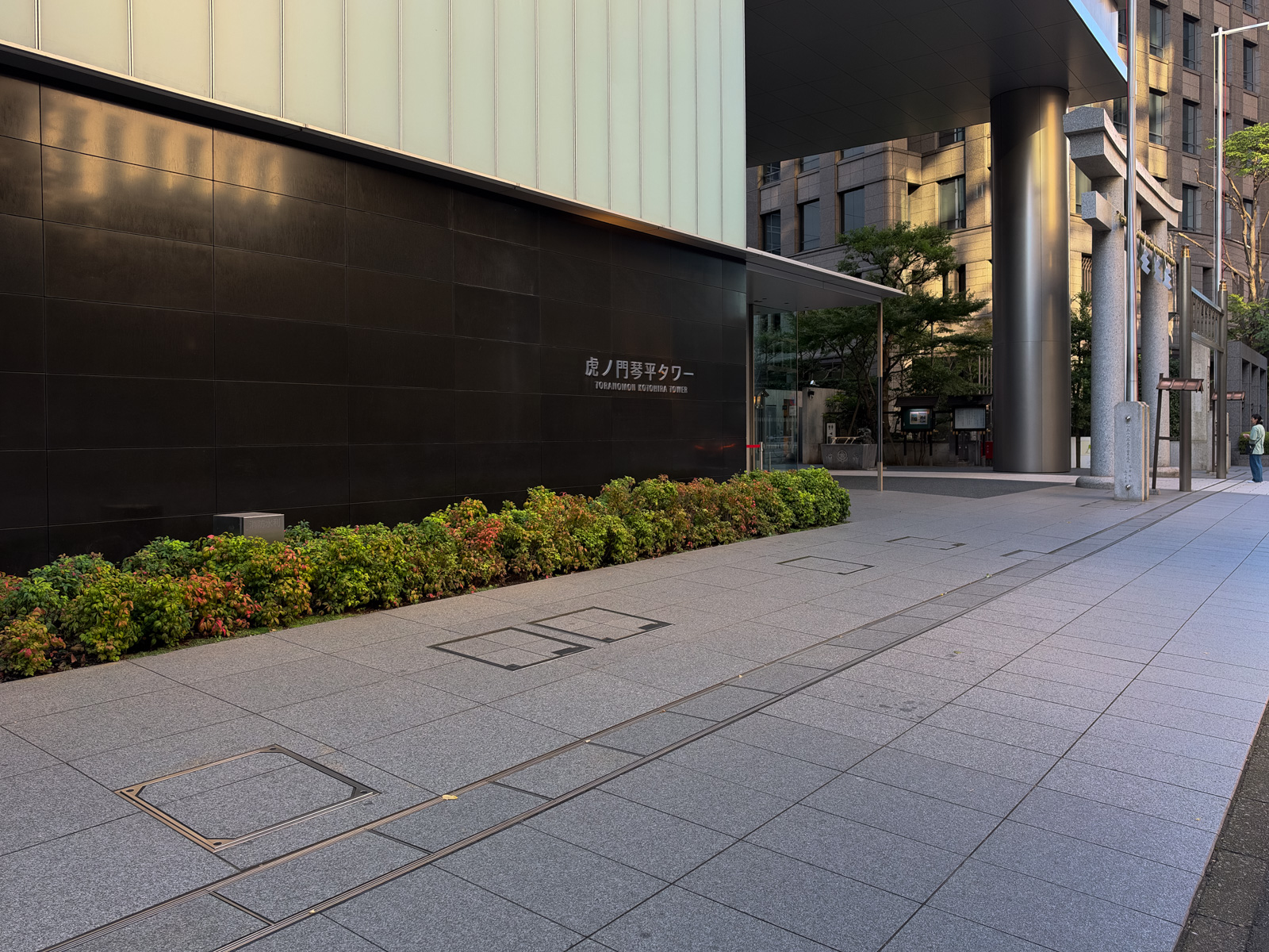 虎ノ門琴平タワー エントランス。「虎ノ門琴平タワー」の看板が黒い壁面に。右奥に鳥居が見える。大庇の下に植栽と石畳