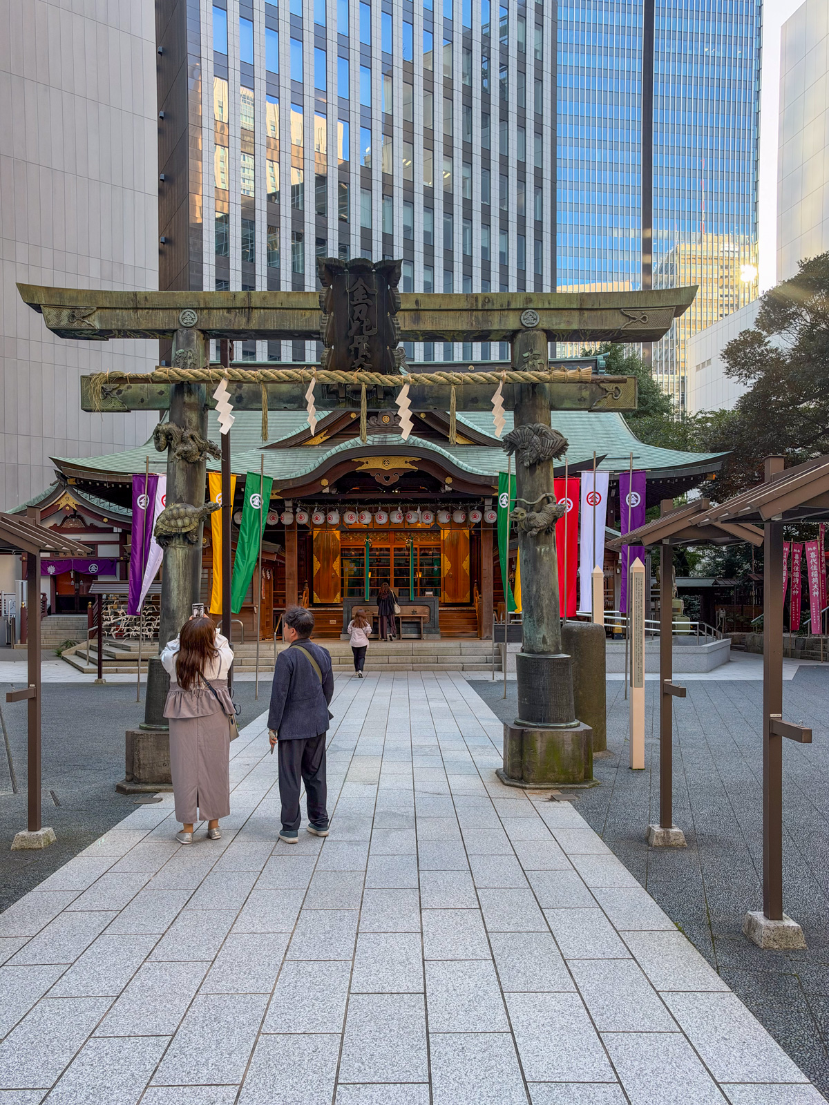 金刀比羅宮 拝殿正面。大きな銅製鳥居、カラフルな幟（紫・黄・緑・赤・白）、社殿。参拝者が二人。背後にタワービル群と空