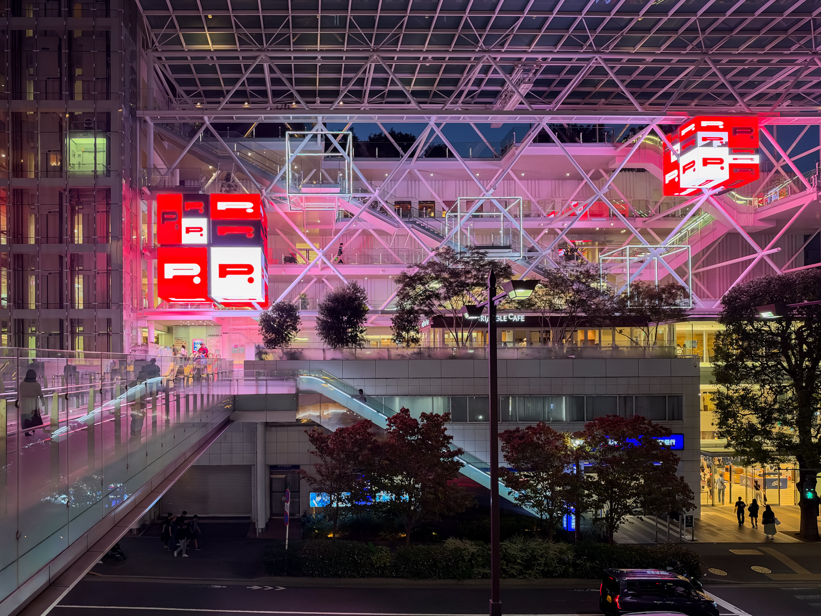 ライズ夜景。白い鉄骨トラスの大屋根の下、赤いLEDサイネージ（R.P.P.P.）がピンク色にライトアップ。RAMBLE CAFEの看板、多層のデッキに人が行き来する