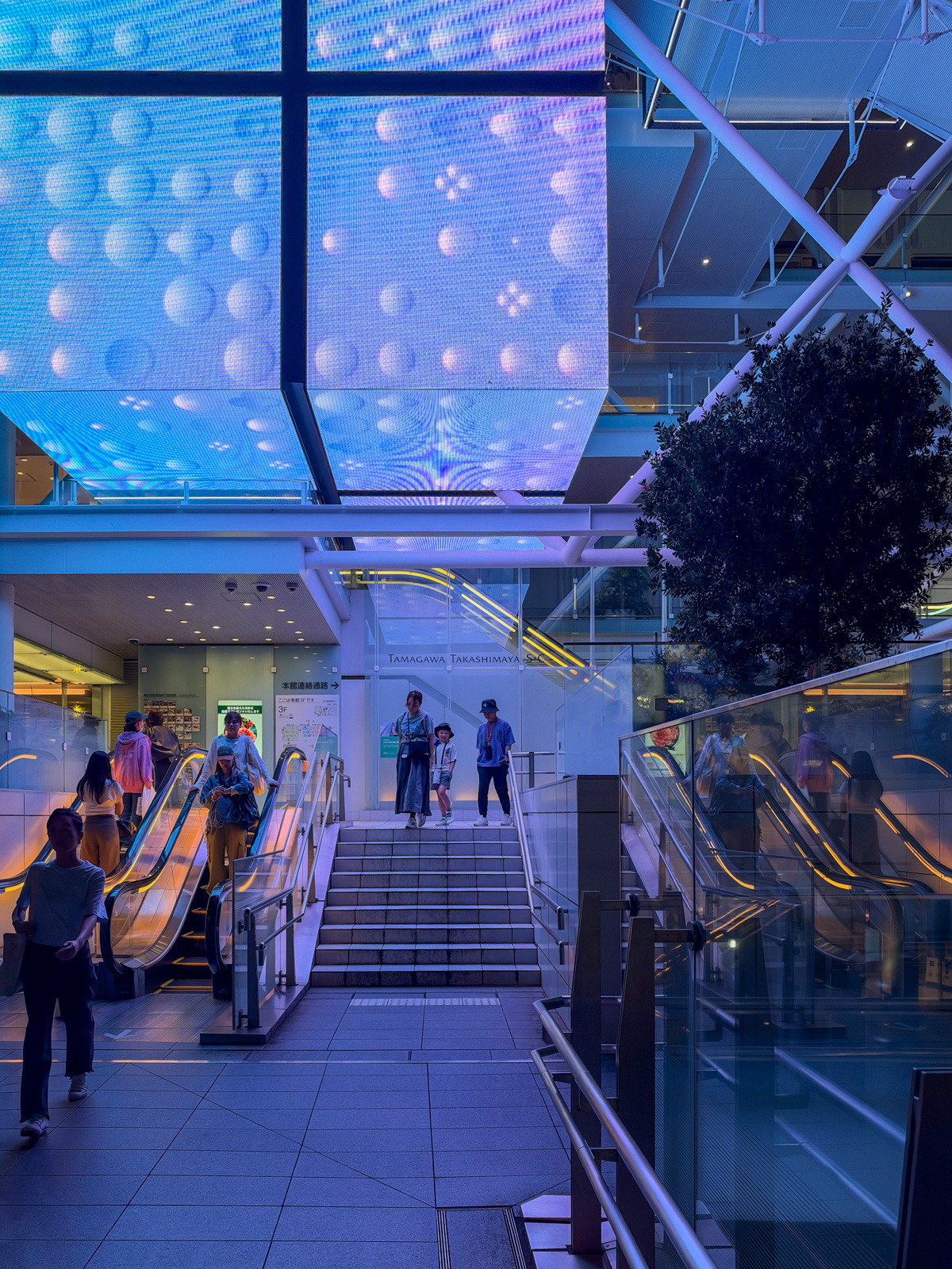 「TAMAGAWA TAKASHIMAYA S.C」の壁面サイン。青いLEDスクリーンに泡のような映像。手前にエスカレーターと階段。人々が下りてくる
