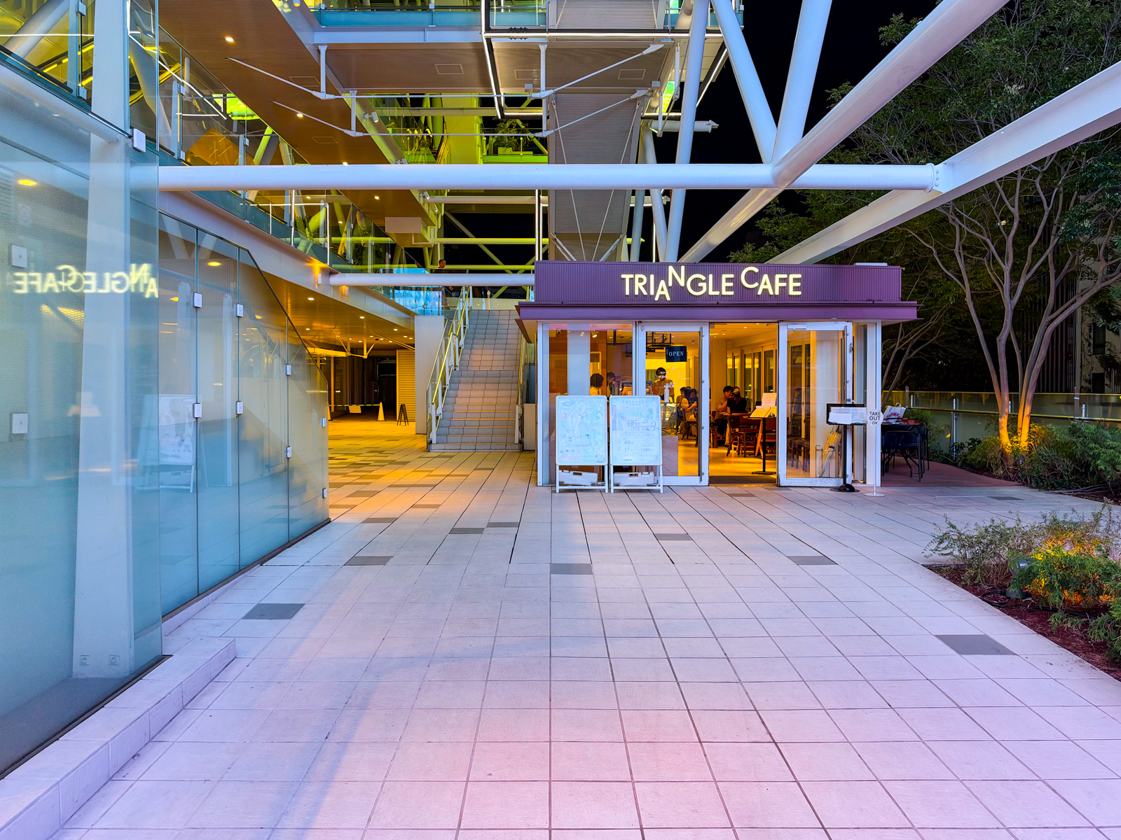 TRIANGLE CAFE。ライズの屋外広場に面した小屋。白いトラス屋根の下、紫色の看板。人が数人座っている。奥にパンケーキ店のガラス壁