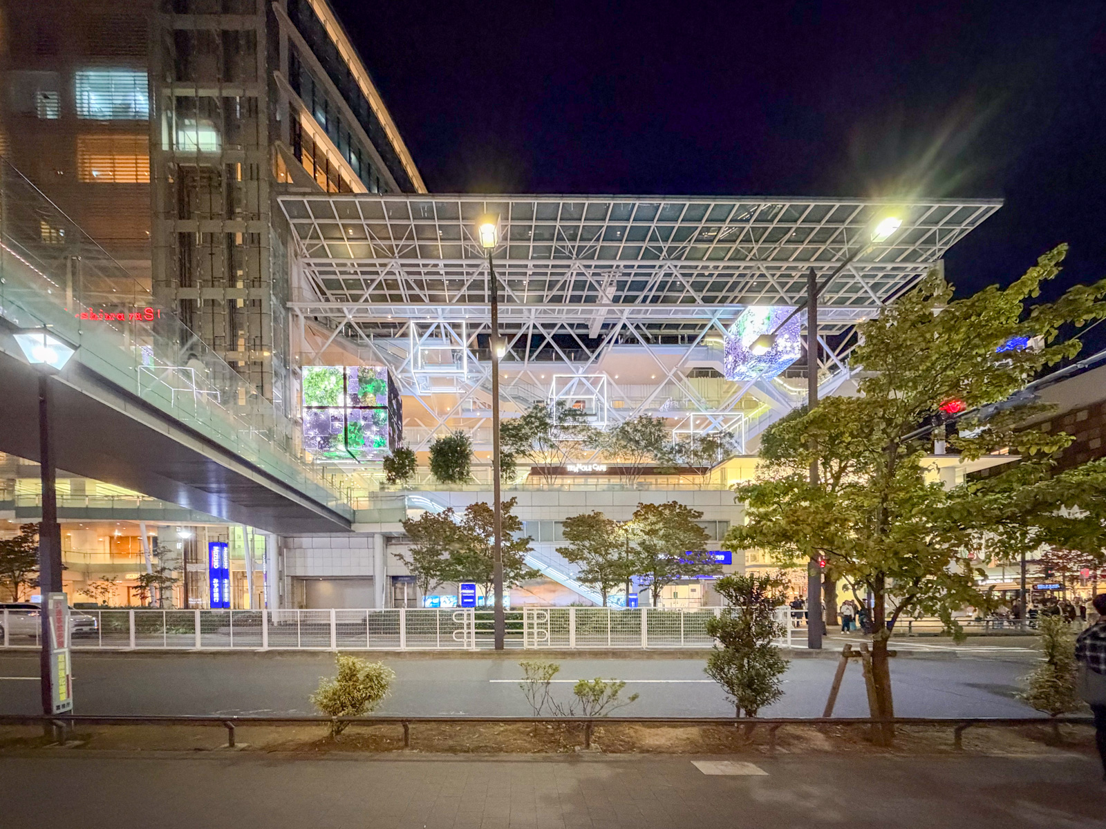 夜のライズ全景。白い鉄骨トラスの大屋根が広がる。左にStarbucksの看板、中央にデジタルサイネージ（緑の樹木）、右に多摩川方向への眺め。歩道から仰ぎ見る